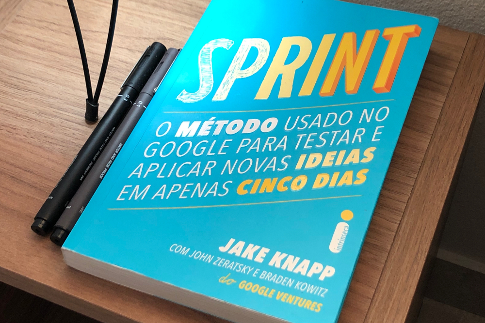 Livro Sprint colocado sobre uma cabeceira de madeira e duas canetas ao lado